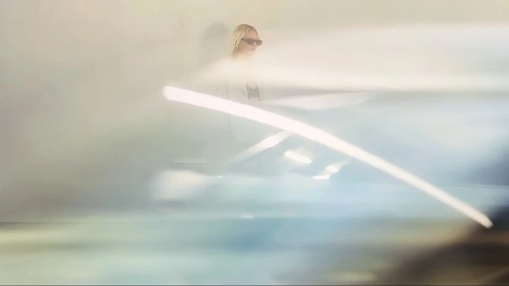 Blonde woman in sunglasses standing behind a blurred foreground element captured by Robbie Mailer-Howat