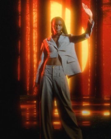 A Black fashion model in a light grey suit with a cropped jacket, standing in a dark space with warm red and orange lighting.