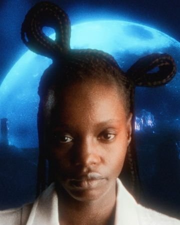 Close-up of an African American woman with braided hair styled with loops, posing in front of a blue celestial background.