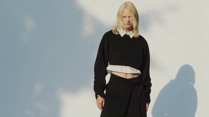 Fashionable adult with long blonde hair wearing a cropped black sweater and skirt in front of a light gray wall with shadows.