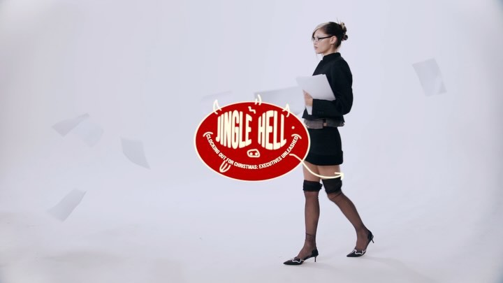 An adult female walks in a bright studio, wearing a black skirt suit, stockings, and heels, holding papers next to a Jingle Hell graphic.
