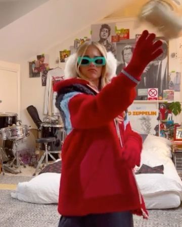 Blonde woman in a red faux fur coat with blue accents, earmuffs, and shades indoors, holding up a hand, with musical instruments nearby.