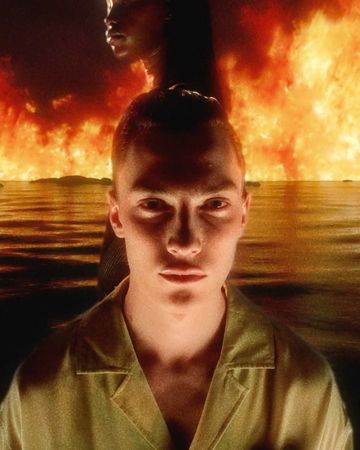 Portrait of a male with short red hair, set against a backdrop of a blurry figure and fiery landscape, creating a surreal visual.