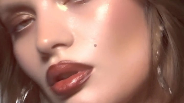 Close-up of a woman's face with glossy red lips, dewy skin, and shimmering gold eyeshadow in a dimly lit, blurred background.