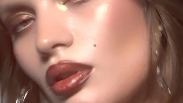 Close-up of a woman's face with glossy red lips, dewy skin, and shimmering gold eyeshadow in a dimly lit, blurred background.
