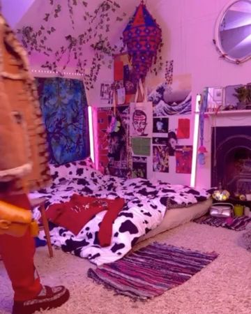 Indoor scene with a bed covered in a cow-print blanket, posters on the wall, and a person standing nearby, bathed in pink lighting.