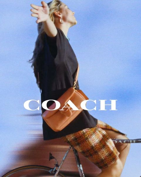 Woman riding a bicycle outdoors wearing a black t-shirt and plaid skirt while carrying a brown leather shoulder bag.