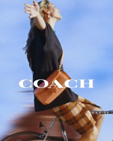 Woman riding a bicycle outdoors wearing a black t-shirt and plaid skirt while carrying a brown leather shoulder bag.