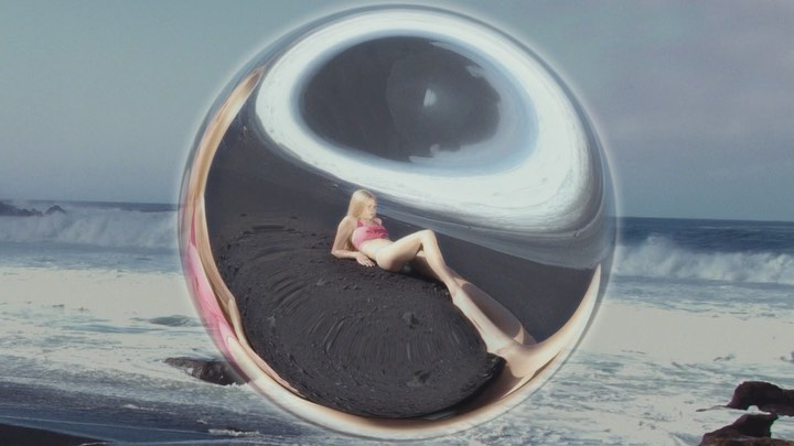 Young woman in pink bikini lying on black sand beach, distorted view through a glass sphere, ocean waves in background.