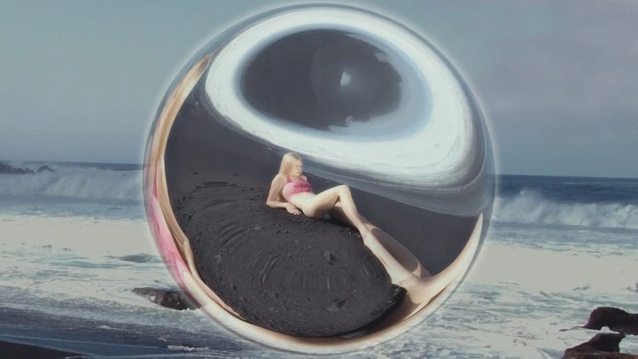 Young woman in pink bikini lying on black sand beach, distorted view through a glass sphere, ocean waves in background.