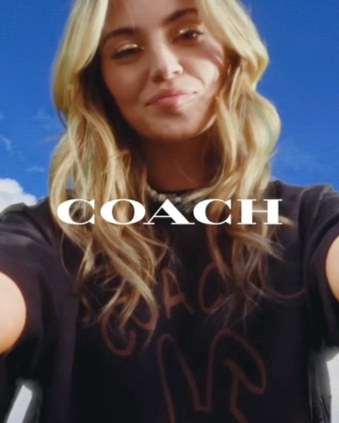 Close-up of a blonde woman wearing a Coach-branded t-shirt against a vibrant blue sky backdrop, creating a fashion-forward outdoor selfie.