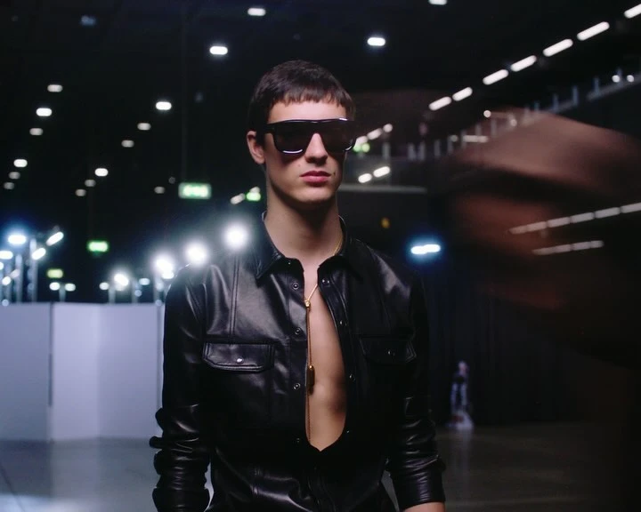 A stylish young adult man with dark hair and sunglasses wears an unbuttoned leather shirt and gold necklace, posing in a dimly lit indoor space.