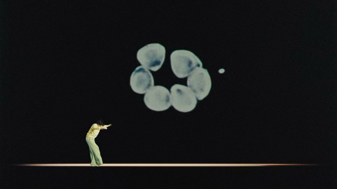 Person in flared pants stands in a dance pose on a stage, illuminated by a spotlight, with an abstract geometric graphic behind them.