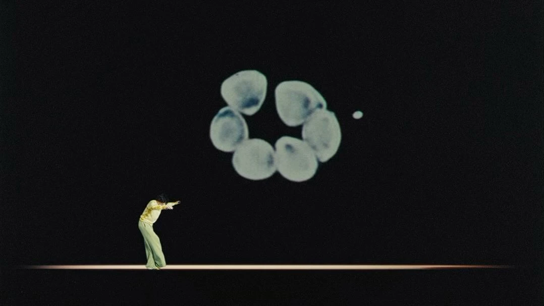 Person in flared pants stands in a dance pose on a stage, illuminated by a spotlight, with an abstract geometric graphic behind them.