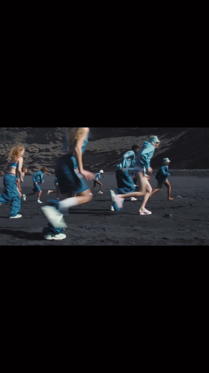 Group of people running on a black sand beach near a rocky hill, captured in motion with blurred legs and sporty fashion attire.