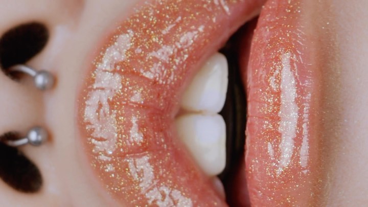 Macro shot featuring an adult female's shiny, pink-toned lips with white teeth visible, and pierced nostril, with a shallow depth of field.