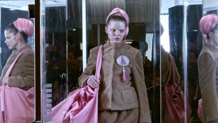 Model wears a brown suit and pink headscarf, carrying a pink bag, standing indoors with reflections, fashion, portrait photography.