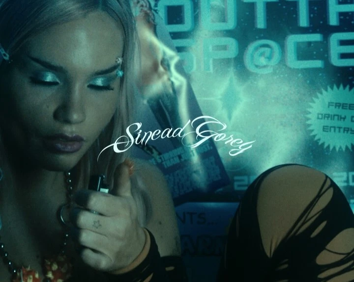 Woman with creative makeup and hair accessories poses in a studio with event poster background, holding a lighter in a close-up.