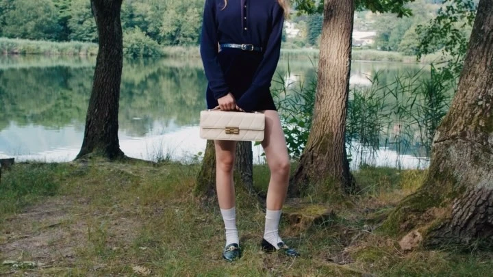 Woman standing outdoors by a lake wearing a navy top, white socks, and loafers while holding a quilted cream handbag.