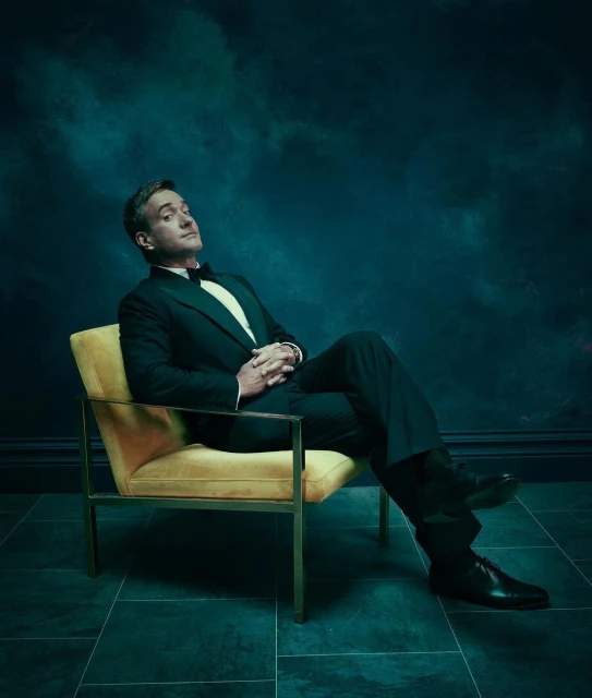 Full-length portrait of a man in a black tuxedo sitting in a yellow chair against a dark teal cloudy backdrop in a photo studio.