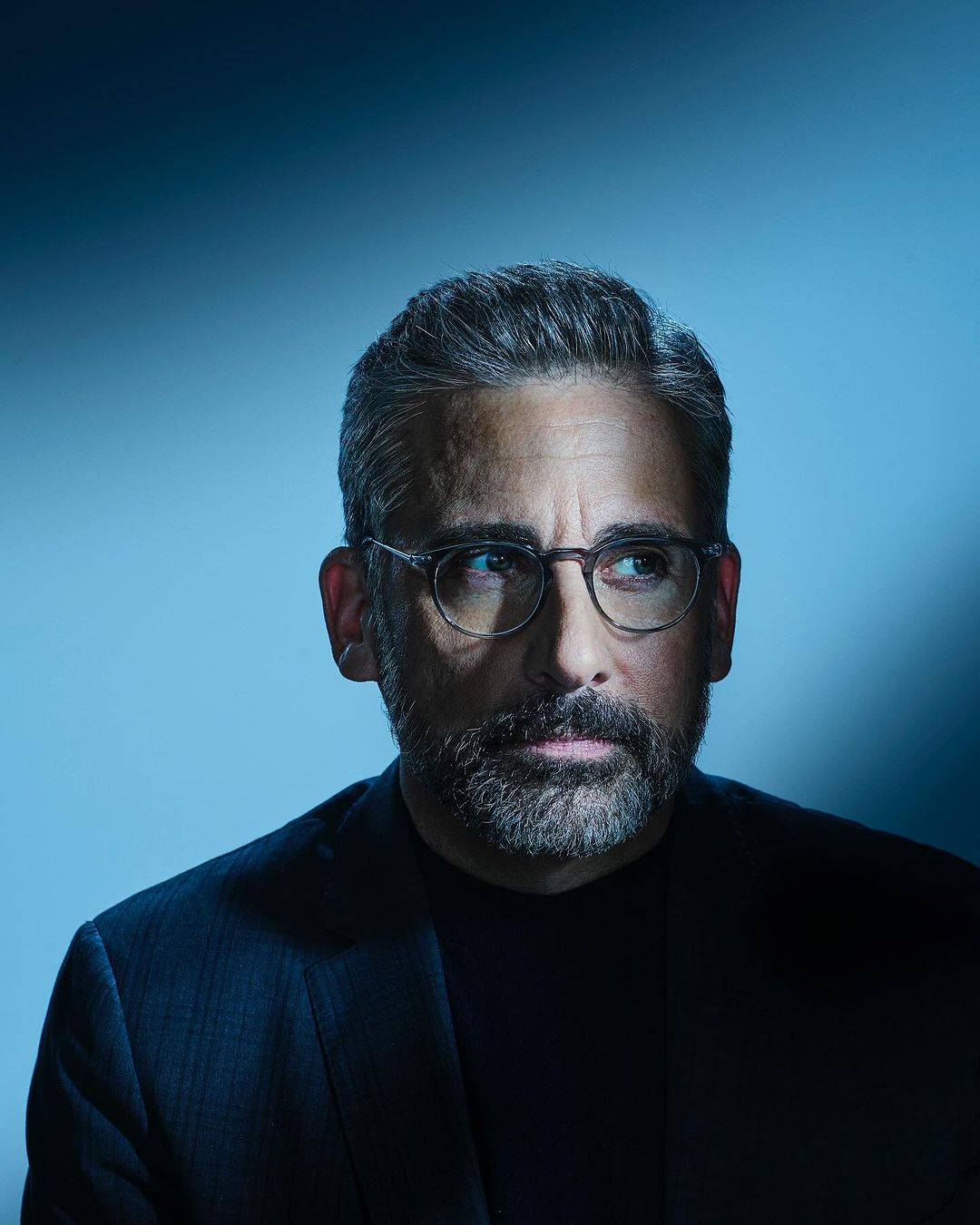 Portrait of a man with graying hair and a beard, wearing glasses and a dark suit, against a gradient blue background in a studio setting.