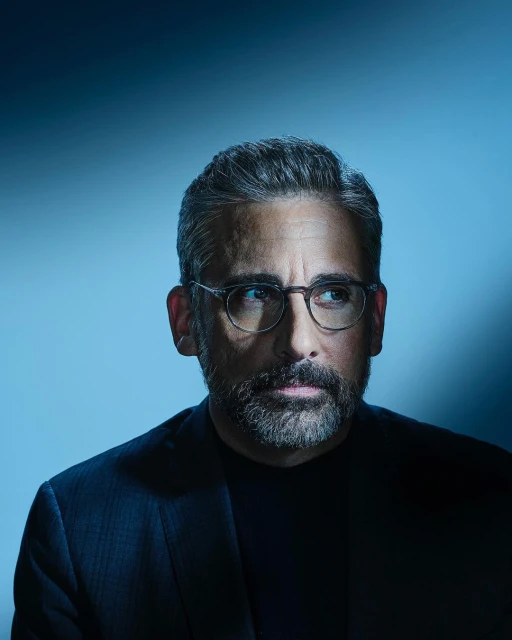 Portrait of a man with graying hair and a beard, wearing glasses and a dark suit, against a gradient blue background in a studio setting.