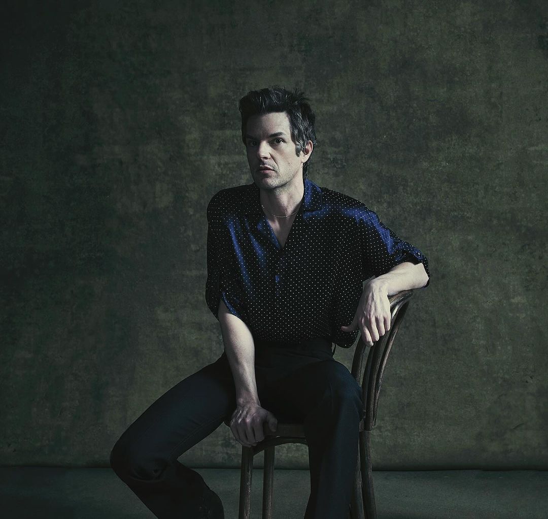 Portrait of a fair-skinned adult male in a dark blue, polka dot shirt, sitting on a chair against a dark, mottled background.