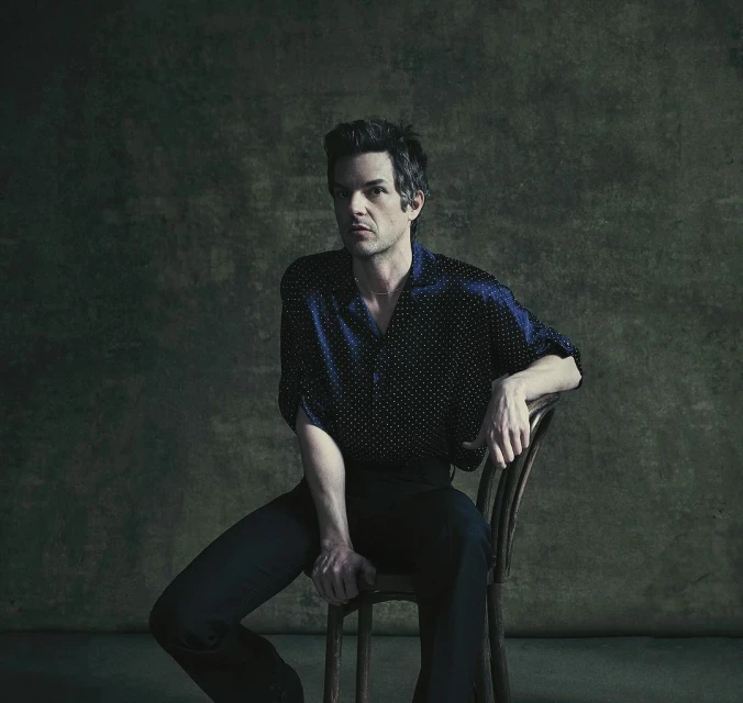 Portrait of a fair-skinned adult male in a dark blue, polka dot shirt, sitting on a chair against a dark, mottled background.