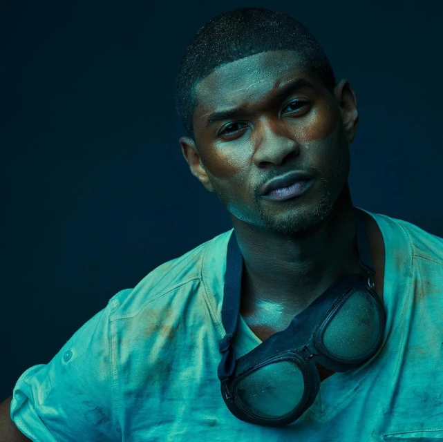 Close-up portrait of a man wearing a t-shirt with goggles hanging around his neck against a dark blue background.