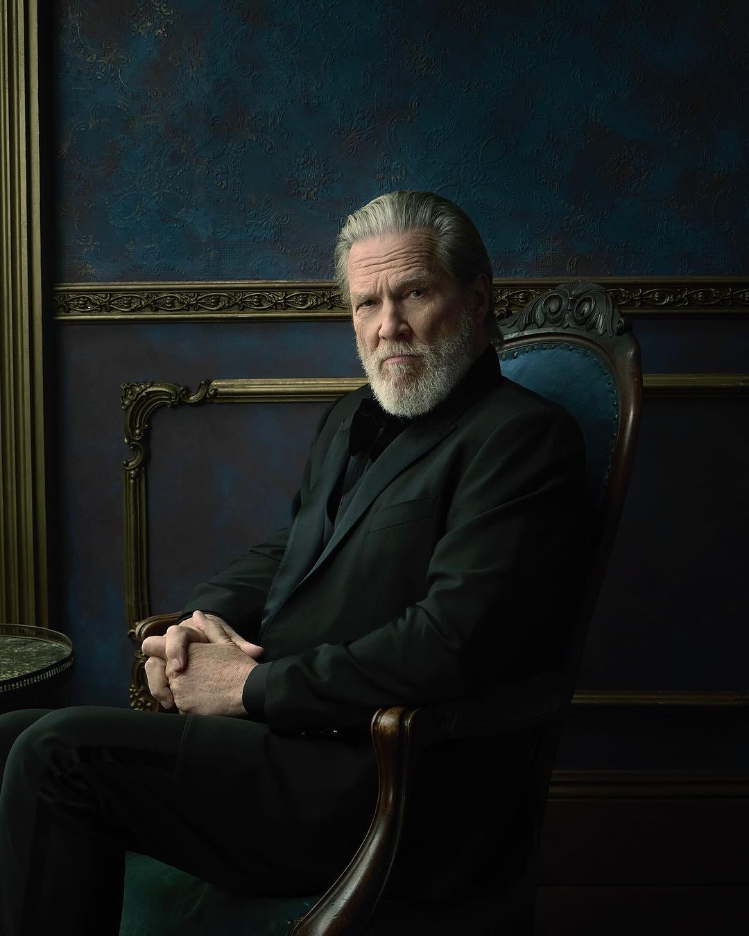 Jeff Bridges seated in an ornate dark wooden chair wearing a black suit jacket against a textured dark blue wall.