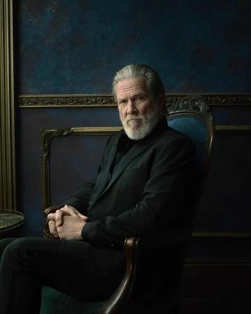 Jeff Bridges seated in an ornate dark wooden chair wearing a black suit jacket against a textured dark blue wall.