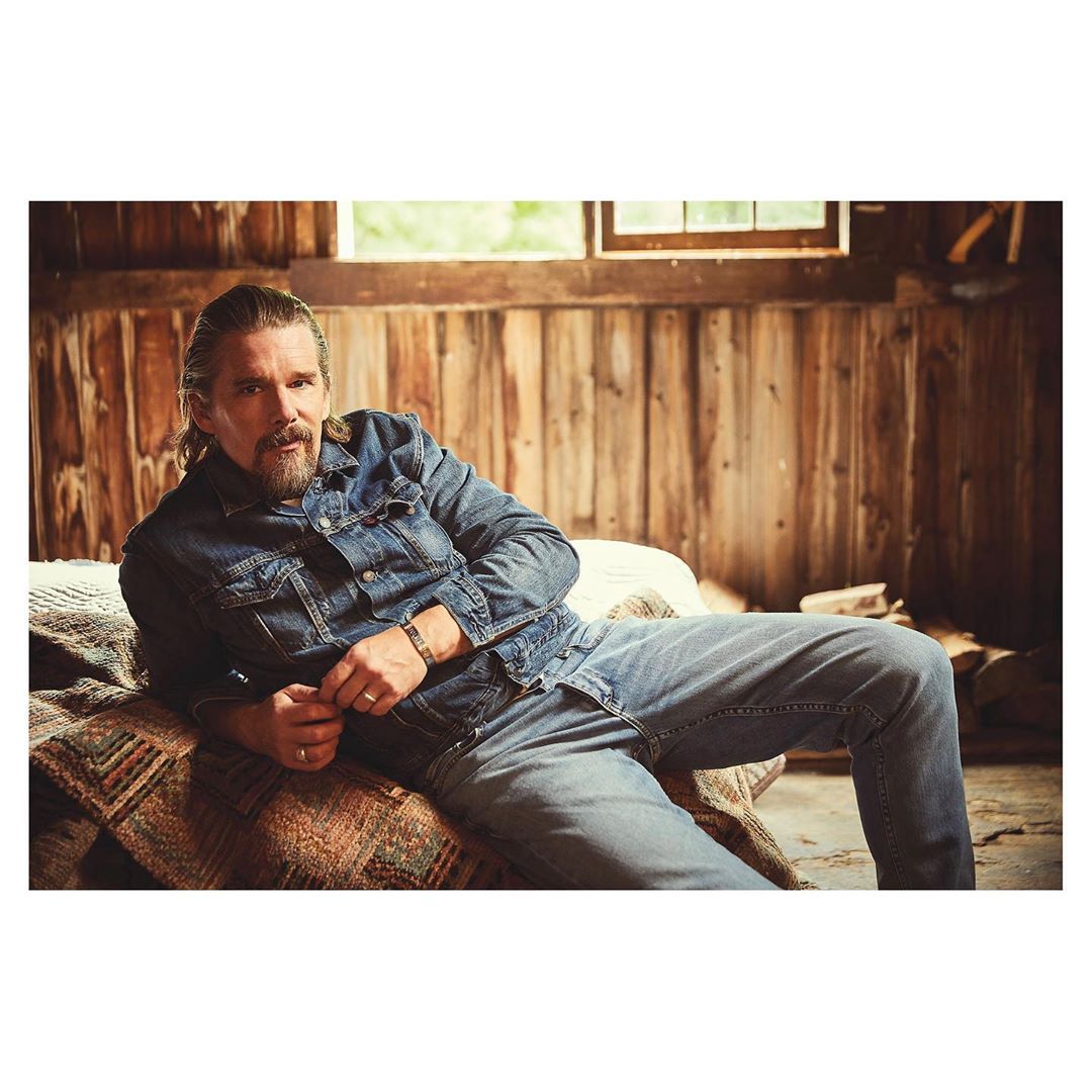 A man in a denim jacket and jeans sits on a patterned bedspread in a wood-paneled room with a window, looking directly at the camera.