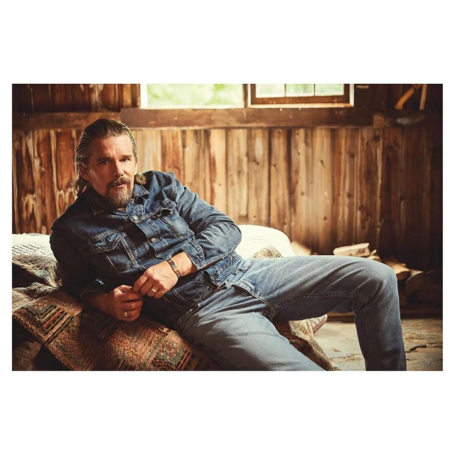 A man in a denim jacket and jeans sits on a patterned bedspread in a wood-paneled room with a window, looking directly at the camera.