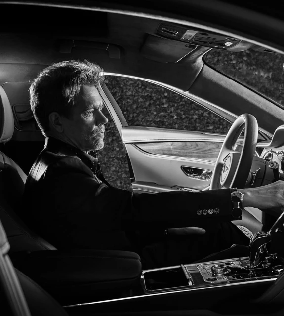 Monochrome shot of a mature man in a suit sitting in the driver's seat of a luxury car with leafy green foliage visible outside.