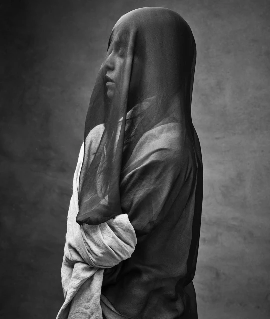 Monochrome portrait of a woman with a draped black veil, her eyes closed, in front of a textured grey background in a studio setting.