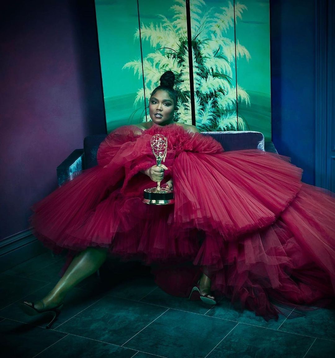 Lizzo sitting in a dark teal room, wearing a large red tulle dress and holding an Emmy award, with a vibrant palm tree painting in the background.