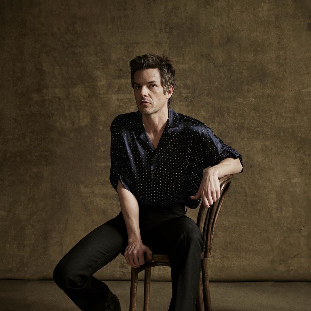 Portrait of a man with dark hair wearing a navy polka dot shirt sitting on a wooden chair in front of a mottled background.