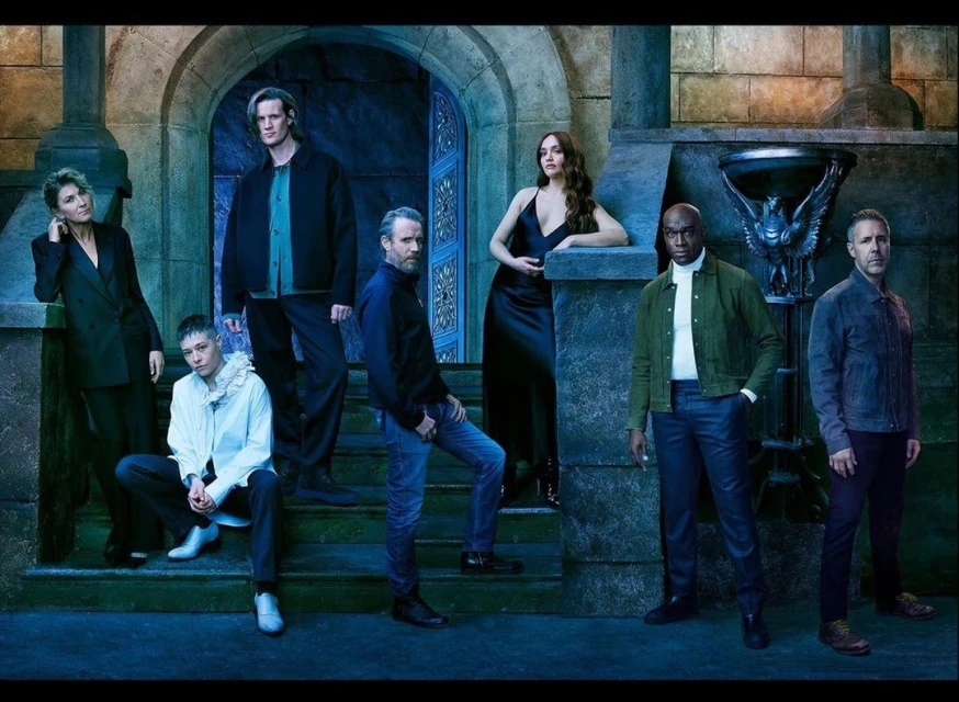 Group portrait of seven actors posing on stone steps with an ornate stone archway in the background, featuring a blue color scheme.