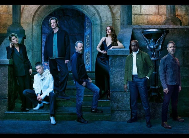Group portrait of seven actors posing on stone steps with an ornate stone archway in the background, featuring a blue color scheme.