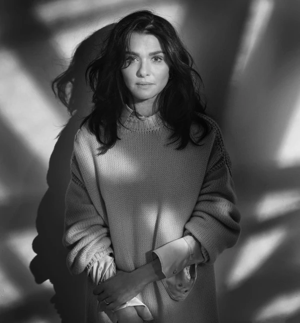 Black and white portrait of a woman in a sweater with patterned shadows on the wall. The woman gazes directly at the viewer.