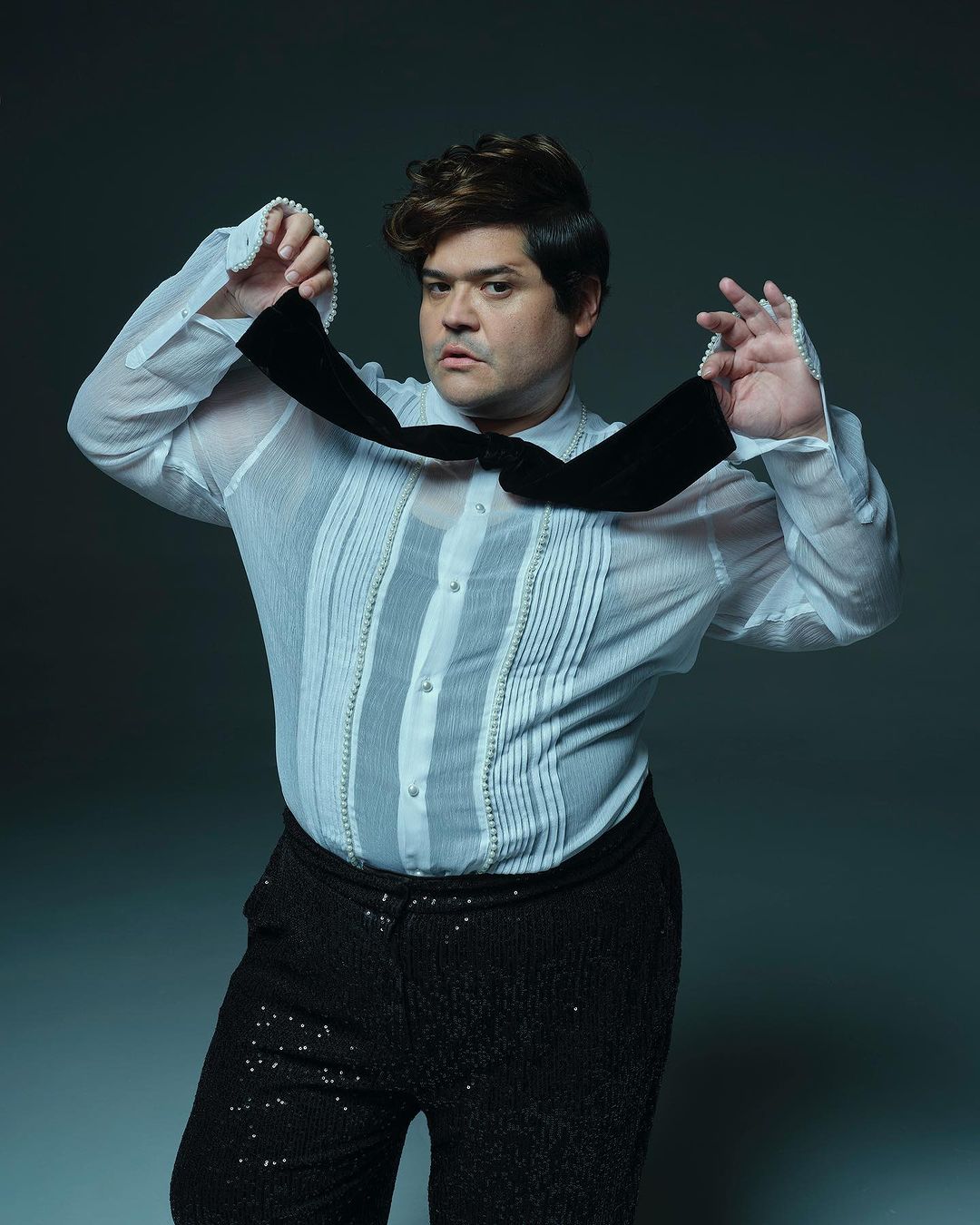 Portrait of a man in a sheer white shirt and shiny black pants adjusting a black bowtie against a gradient grey backdrop.