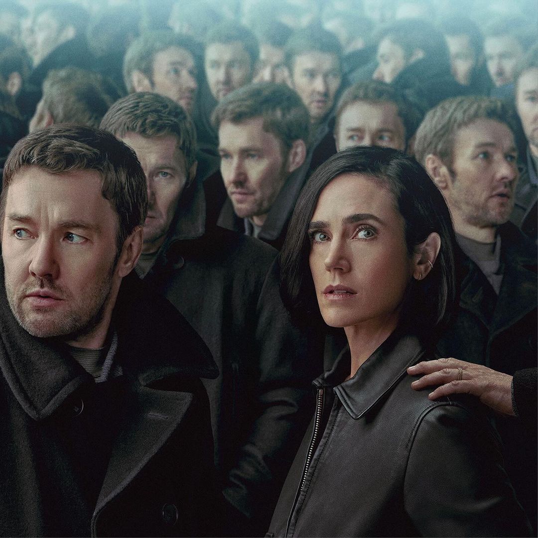 Jennifer Connelly and male actor in a crowd, dressed in dark coats, looking with serious expressions, indoor lighting, overcast mood.