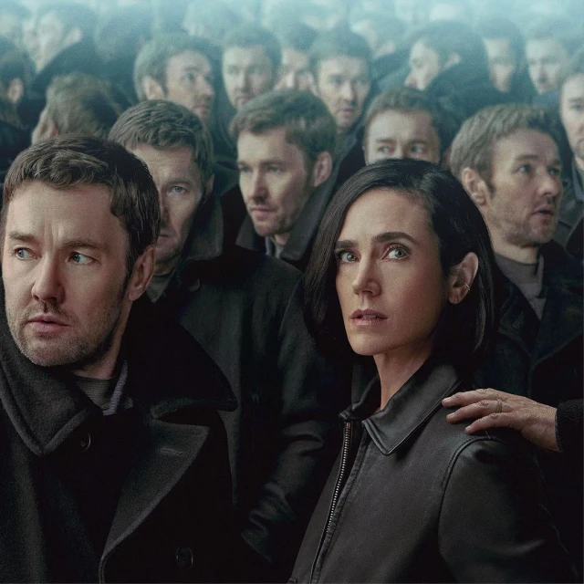 Jennifer Connelly and male actor in a crowd, dressed in dark coats, looking with serious expressions, indoor lighting, overcast mood.