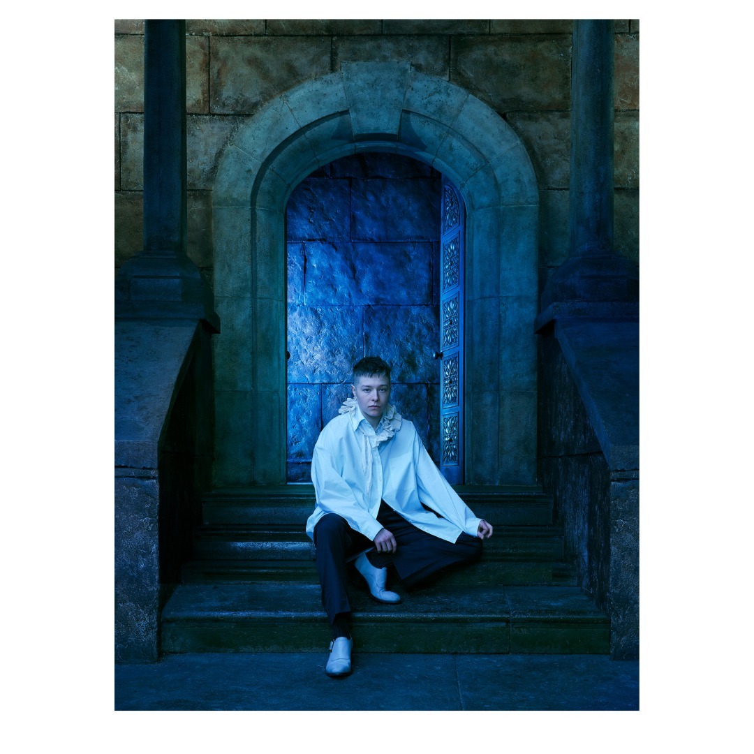 Portrait of a person sitting on stone steps in front of a stone doorway and blue doors in an indoor, castle-like environment.