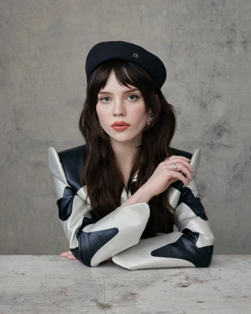 Portrait of a woman with dark hair wearing a black beret and a silver and black structured jacket against a textured wall.