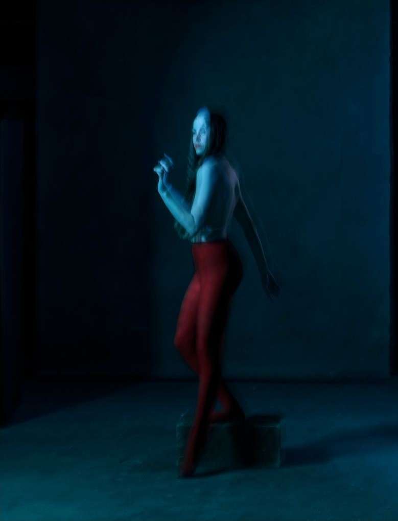 Adult woman in red leggings standing on a box in a dimly lit interior space, with blue light casting shadows across the scene.