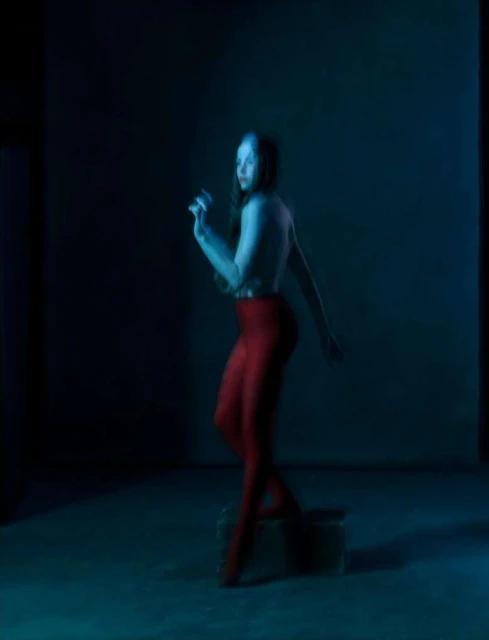 Adult woman in red leggings standing on a box in a dimly lit interior space, with blue light casting shadows across the scene.
