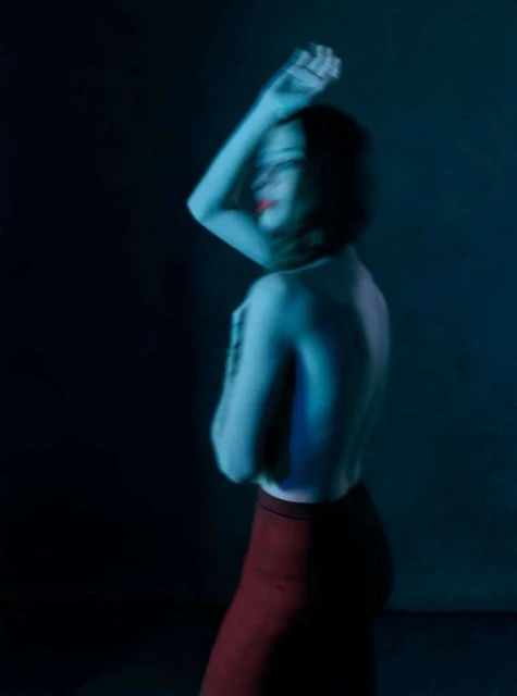 Backlit nude woman with red lipstick poses in a dark, low-key studio environment with artistic lighting highlighting her shoulders and neck.