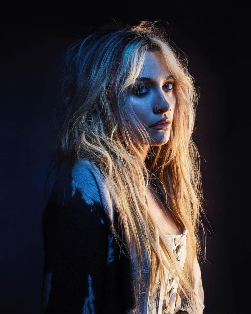 Portrait of a young woman with blonde hair wearing a white top and cardigan in front of a dark background with blue and gold lighting.
