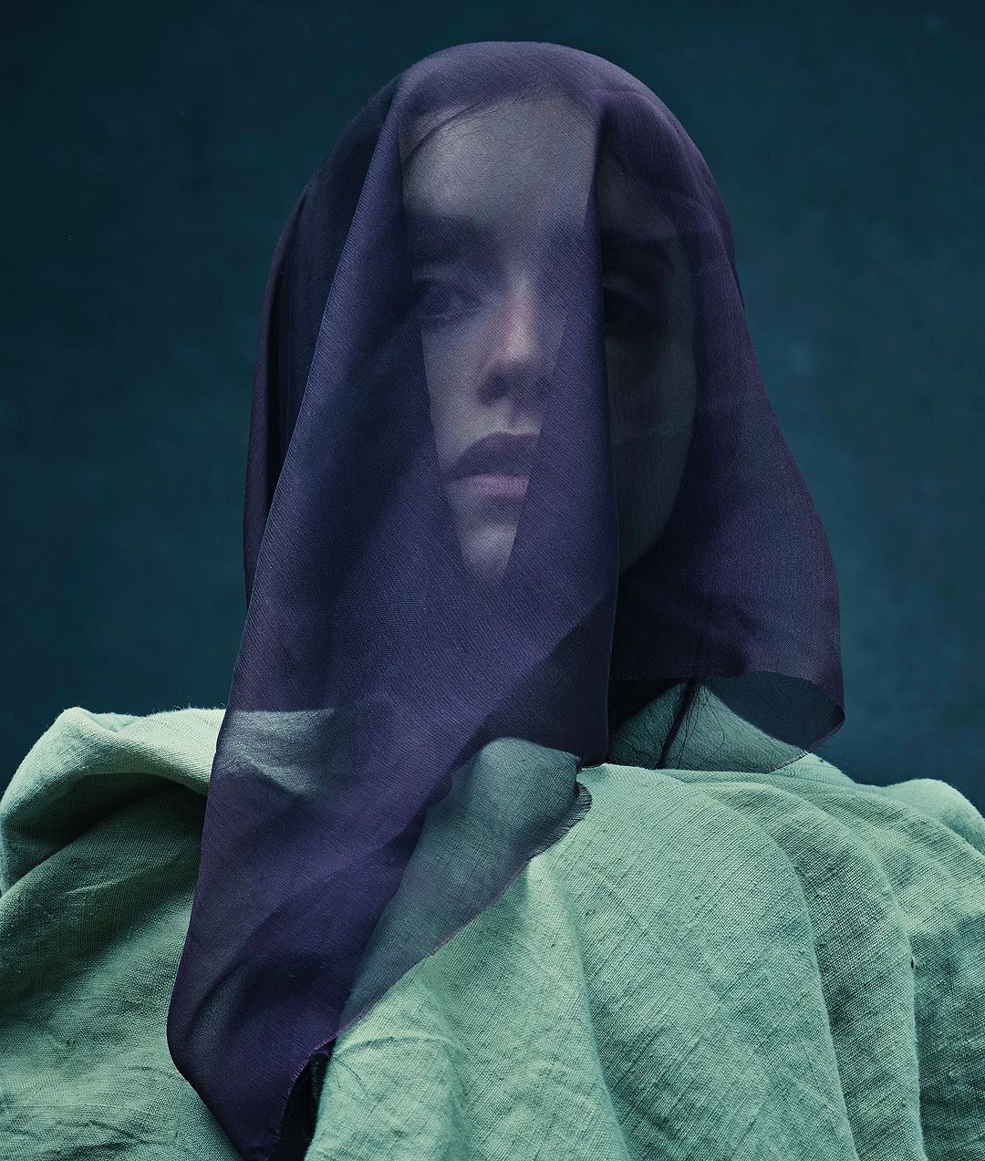 Portrait of a person with a purple sheer veil over their face, wearing a green textured garment against a dark background.